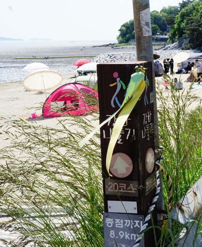 나들길 이정표 말뚝 뒤로 보아는 분홍색 텐트, 흰색 텐트, 그외 여러 텐트들, 모래사장에 여러사람들, 멀리 드넓은 갯벌, 하얀 하늘,