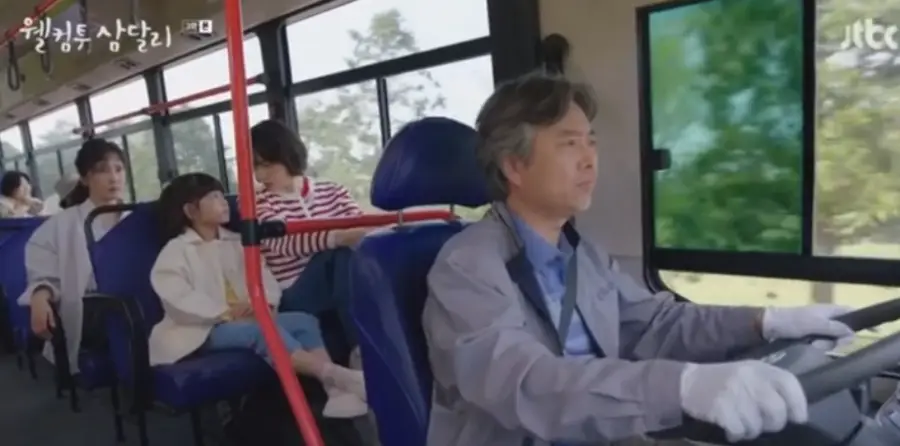 웰컴투삼달리- 머리가 히끗히끗한 아저씨가 파란셔츠를 입고 버스 운전대를 잡고 운전하는 모습