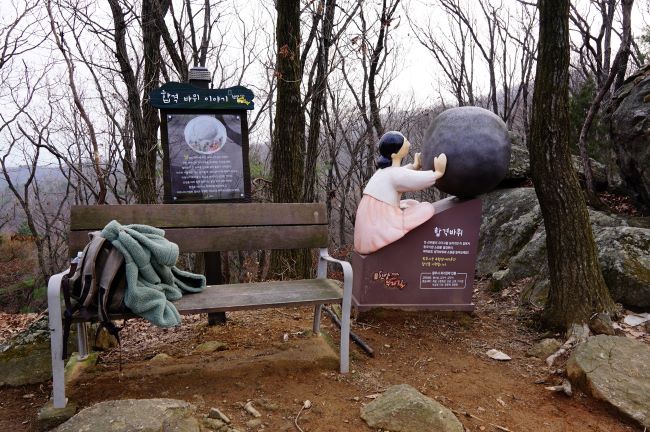 합격바위 앞에 흰옷 입고 앉아 큰 바위를 밀고 있는 어머니 조형물&#44; 왼쪽에 합격바위 안내문&#44; 그 앞 벤치에 내려놓은 배낭&#44; 하얀 하늘&#44;