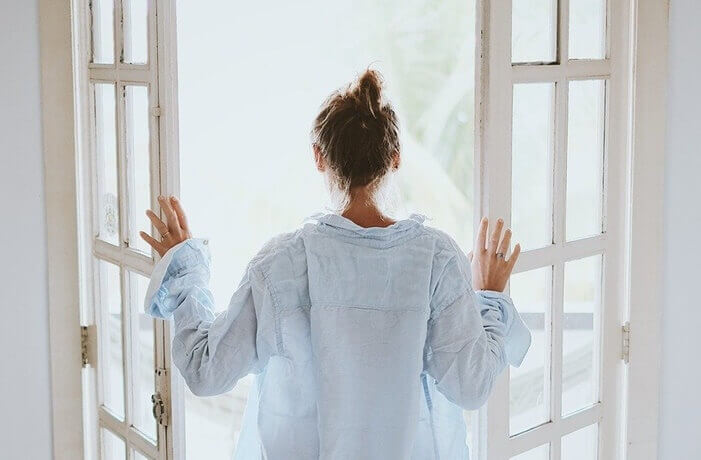 옅은 하늘색의 셔츠를 입은 여자가 하얀 창틀의 창문을 열고 있는 모습