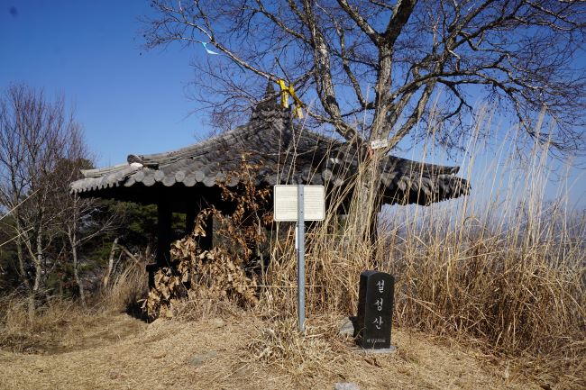 푸른 하늘&#44; 팔각정&#44; 기와지붕&#44; 그 앞에 큰 나무&#44; 그 앞에 검은 돌에 새긴 설성산 정상석&#44; 흰글씨&#44; 정자 앞을 어지럽게 가리고 있는 갈대&#44;