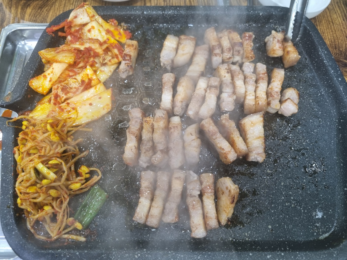 불판위에 삼겹살과 김치 그리고 콩나물이 익어가고 있는 사진. 삼겹살을 먹기 좋게 잘라놓았다.