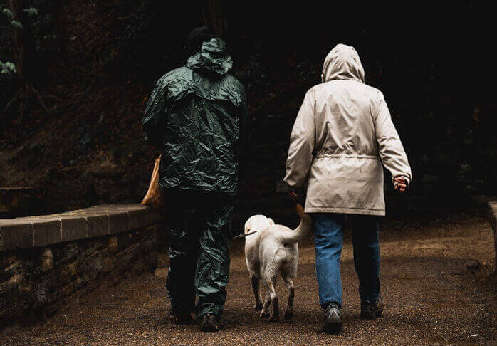 우비를 입은 두사람과 그 사이에 밝은색 털의 개 한마리가 비를 맞으며 걷고 있는 뒷모습