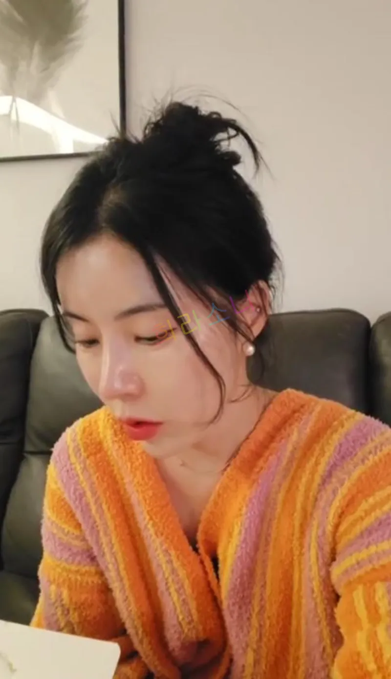 머리를 질끈 묶은 10기 영숙이 같은 오렌지와 핑크 스트라이프 패턴의 가운을 입고 아래를 내려다보며 집중한 표정을 짓고 있다. 그녀는 무언가를 보고 있는 듯하다. 배경에는 검은 가죽 소파가 보인다. 자막에는 &amp;quot;희라소니&amp;quot;라는 텍스트가 보인다.