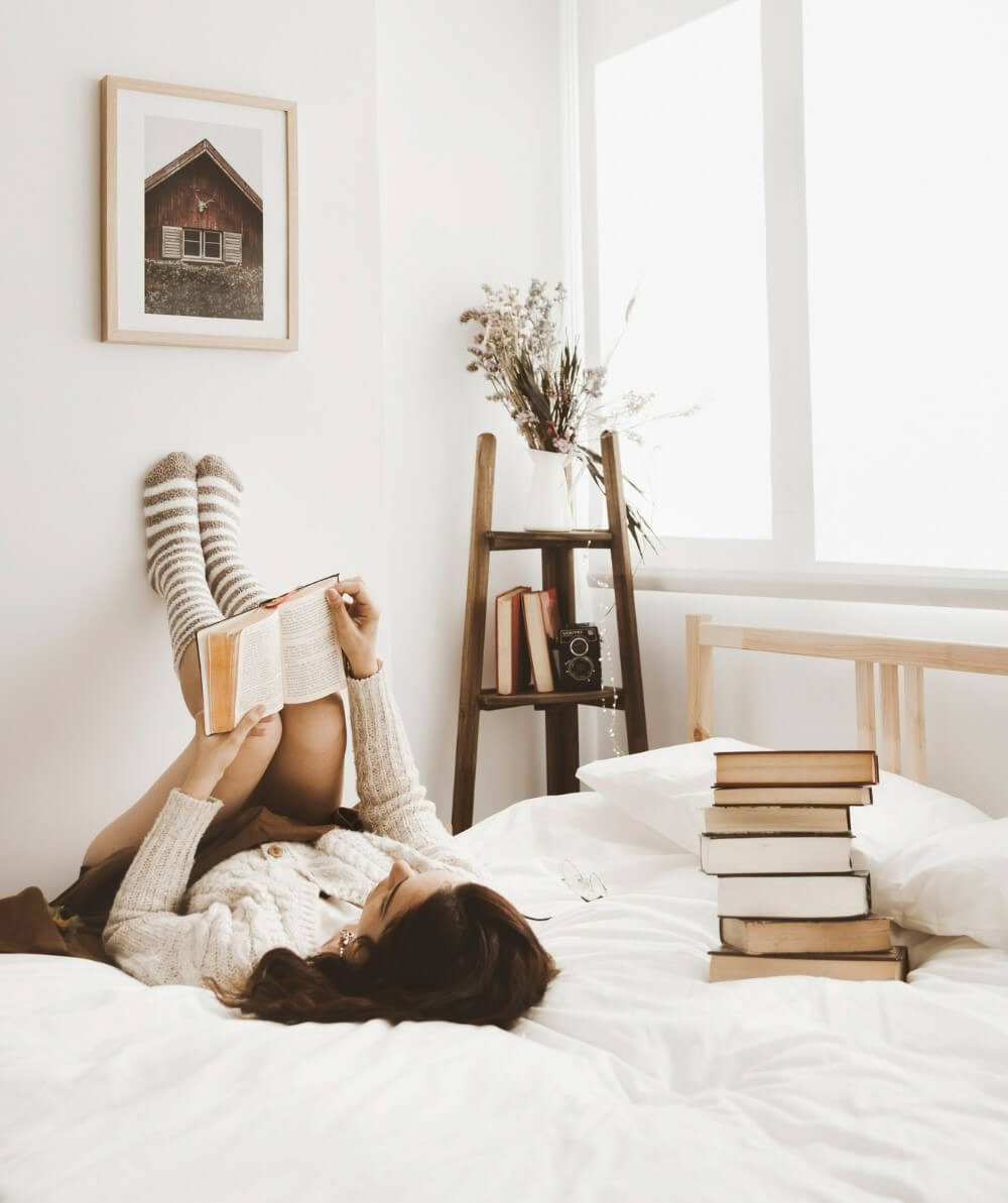 침실 침대에 책을 쌓아놓고 한 여성이 침대에 누워 책을 읽고 있다.