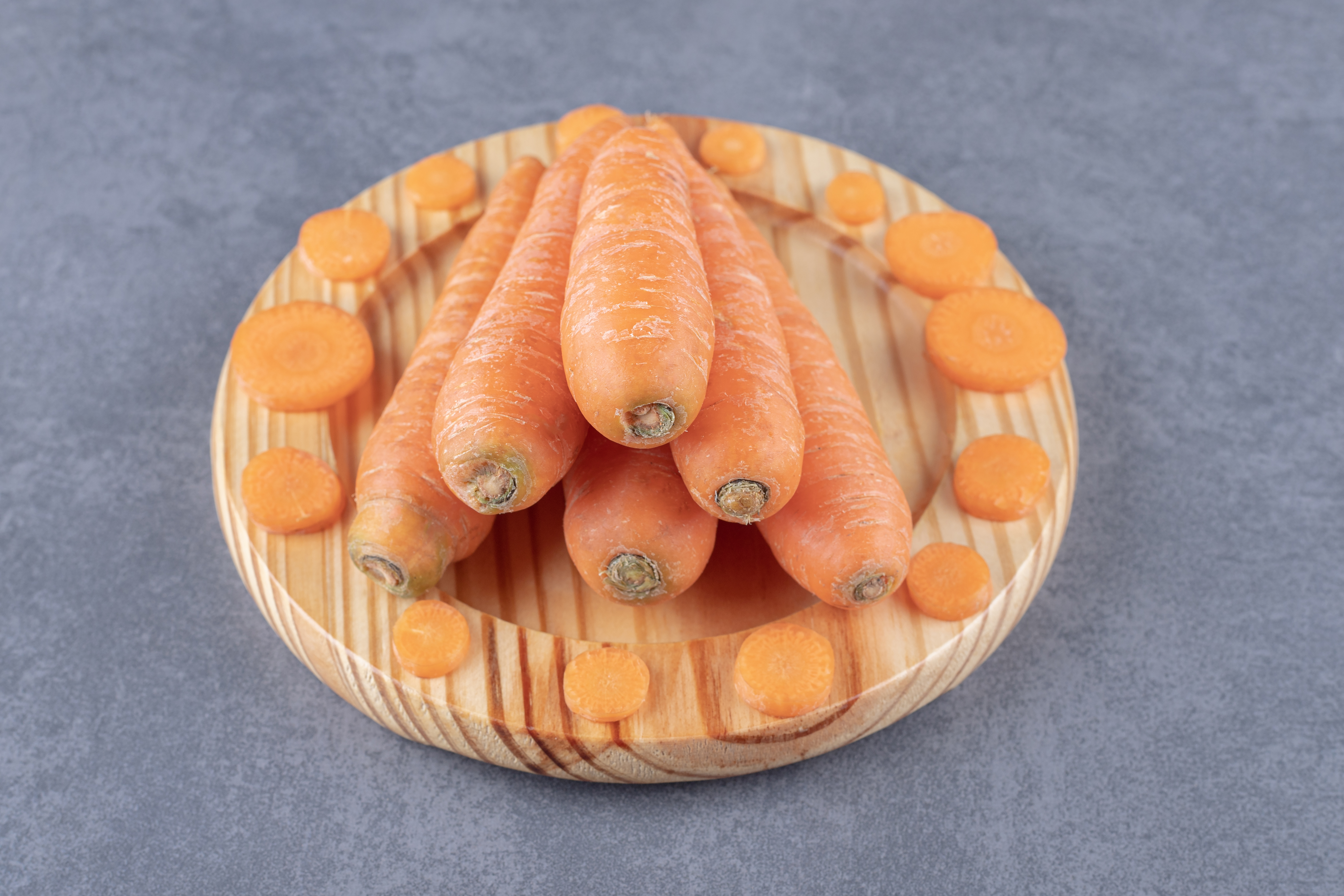 동그란 나무 접시 위에 당근을 6개 가운데 두었고&#44; 접시 테두리 부분에 당근을 얇게 썬 것을 둥그렇게 놓아두고 찍은 사진 (whole-sliced-carrots-wooden-plate-marble-surface)