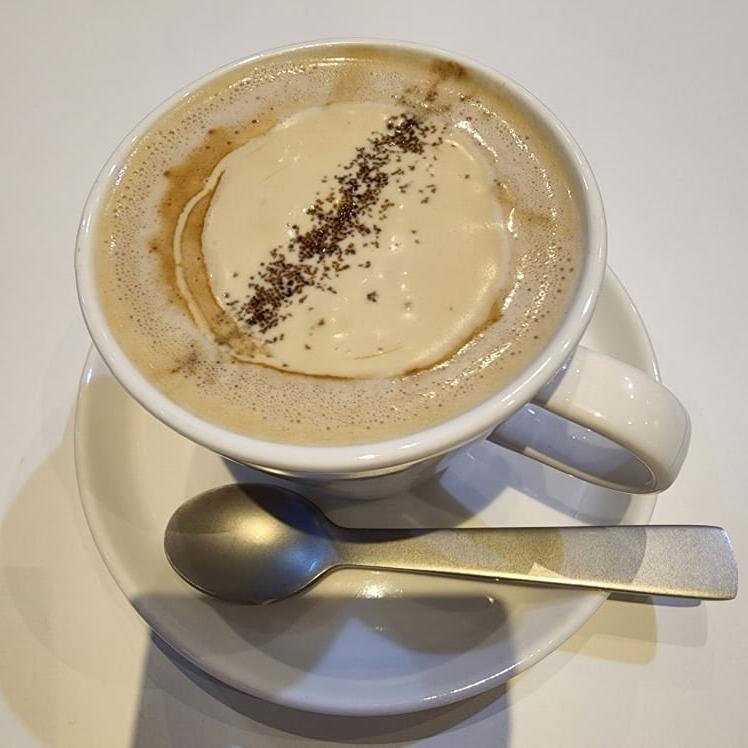 하얀 컵 받침이 있는 찻 잔에 커피 크림이 올려져 있고 커피 가루가 뿌려져 있다. 조그만 스푼이 컵 받침 위에 올려져 있다.