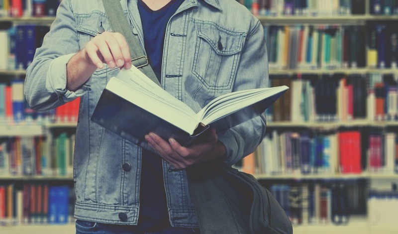 A student holding an open book in a university library, symbolizing the intent to resume academic studies after military service.