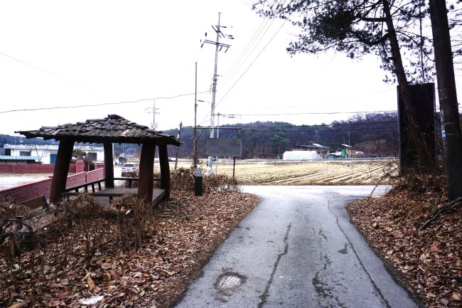 왼쪽에 사각 정자&#44; 우측에 큰 나무&#44; 가운데 농로 T 자형 길&#44; 건너편에 논밭&#44; 흐린 하늘&#44;