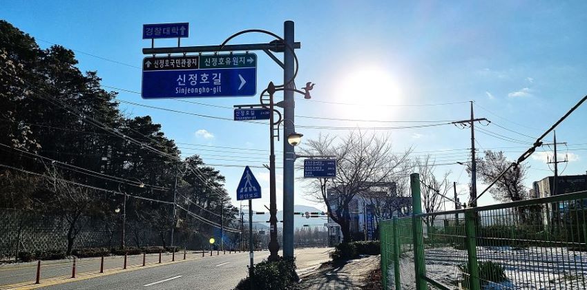 가운데에 우뚝한 교통표지판, 파란바탕에 흰글씨로 쓰인 신정호길과 화살표 흰색, 푸른 하늘, 역광, 왼쪽에 차로(신정로), 우측에 그린 철망,