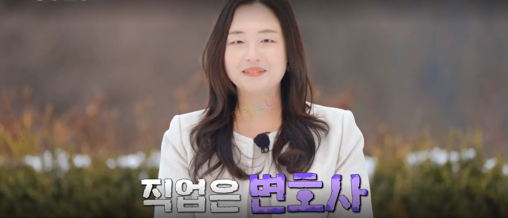같은 여성이 웃으며 클로즈업된 장면. 붉은 립스틱을 바르고 자연스럽게 내린 머리를 유지하고 있으며&amp;#44; 귀걸이를 착용하고 있다. 자막은 &lsquo;직업은 변호사&rsquo;.