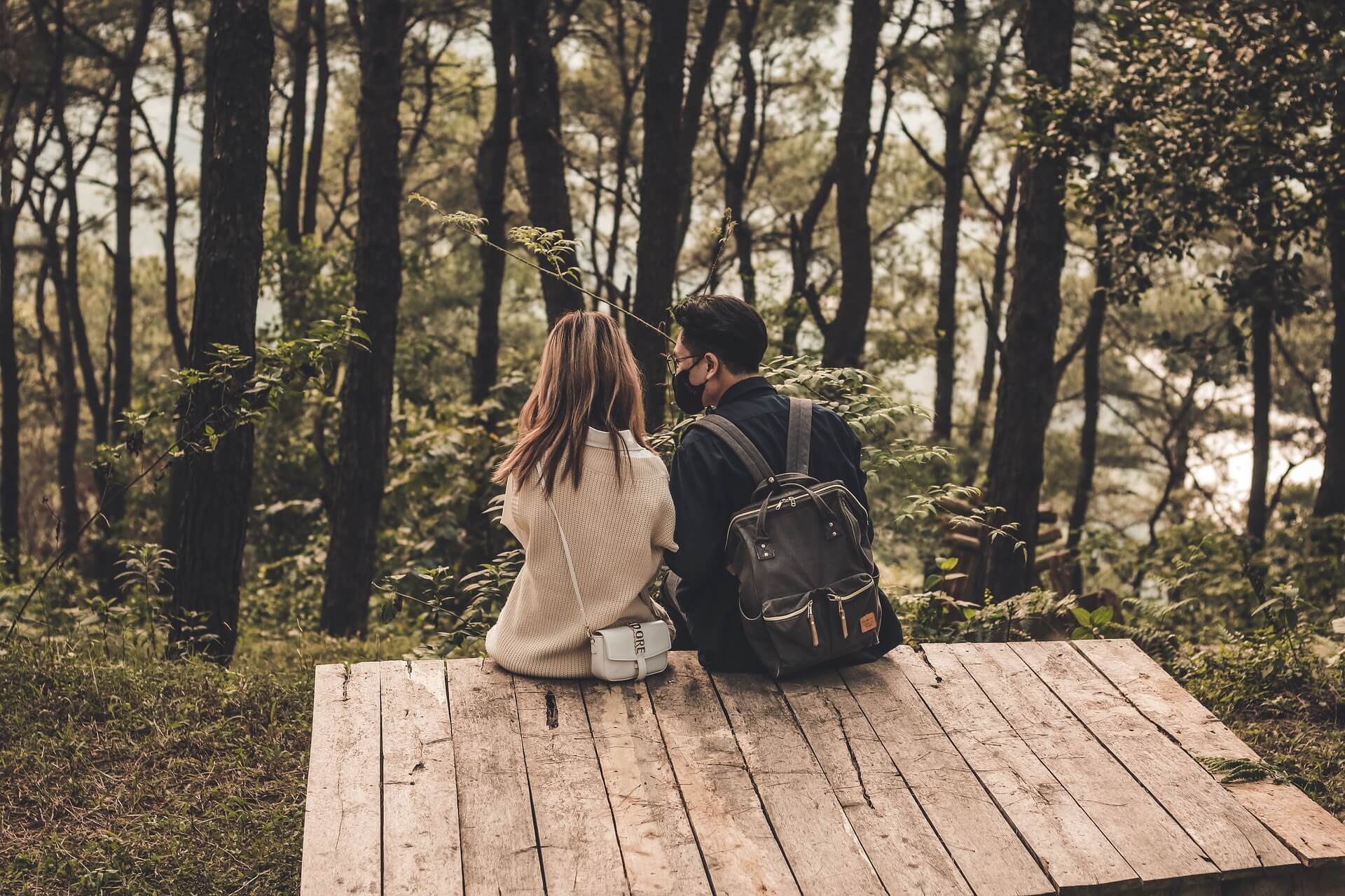 숲 속 나무 데크 위에 앉아 대화를 나누는 백팩을 멘 남성과 크림색 스웨터를 입은 여성의 뒷모습