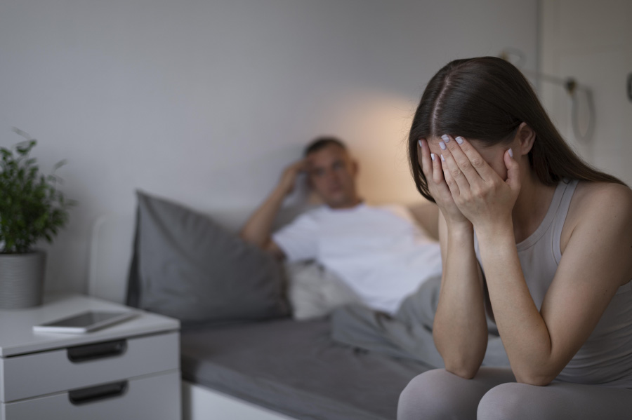 침실의 침대 위에 하얀색 티셔츠를 입은 남성이 침대에 기대어 앉아 있고, 여성은 침대 모서리에 걸터 앉아서 양쪽 손으로 얼굴을 가리며 슬퍼하고 있는 것을 찍은 사진
