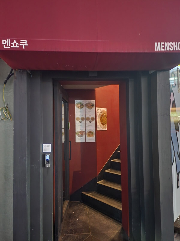 신사역 근처 라멘 맛집 멘쇼쿠의 입구. 매우 좁은 입구에 빨간 차양이 쳐져 있고 왼쪽 귀퉁이에 한글로 &#39;멘쇼쿠&#39;라고 쓰여 있다. 계단이 있는 벽은 모두 붉은색으로 칠해져 있고 라멘 포스터가 잔뜩 붙여져 있다.