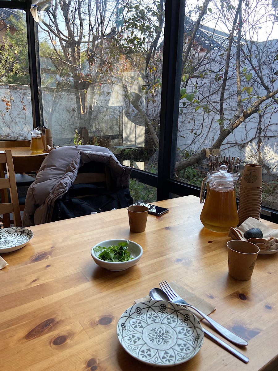 유리창 너머로 작은 정원과 나무가지가 있고 식사준비가 세팅된 테이블