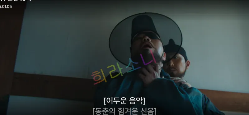 은 갓을 쓴 남자가 뒤에서 지행수의 목을 움켜쥐고 있습니다. 두 사람 모두 긴장된 모습이며&amp;#44; 배경은 나무 기둥이 있는 방 내부입니다. 자막은 &amp;quot;[어두운 음악] [동춘의 힘겨운 신음]&amp;quot;.