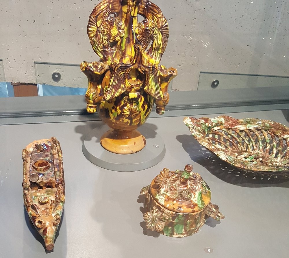 유리 진열장 안에 다양한 크기와 형태의 도자기들이 전시되어 있다. 중앙에는 복잡한 색감의 장식기와 꽃병이 있고, 주변에는 유약 처리된 주전자, 접시, 그릇들이 나란히 배열되어 있다.