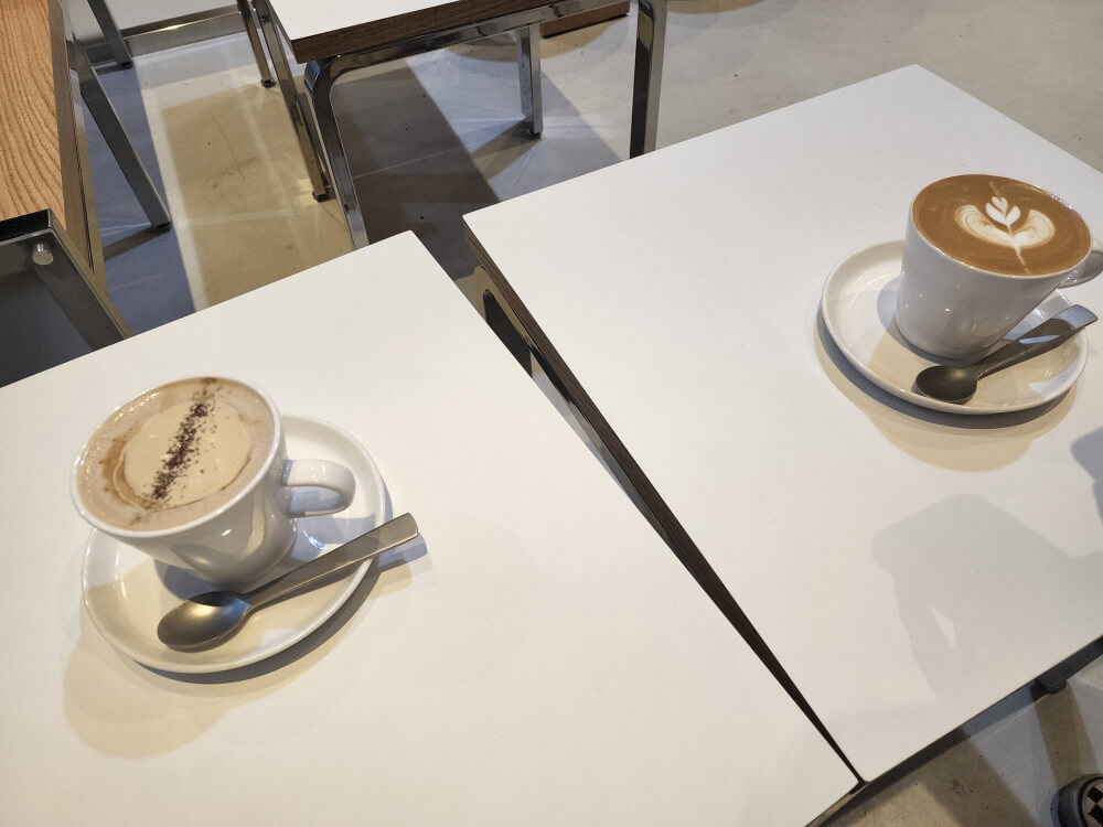 하얀 테이블 위에 코펜하게너와 데니쉬 라떼가 놓여져 있다. 코펜하게너는 하얀 찻잔에 커피 휘핑 크림이 올려져 있고 데니쉬 라떼는 라떼 아트가 되어 있다.