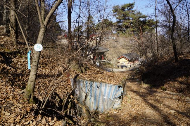 산 아래로 내려가는 산길&#44; 산 아래 보이는 사찰 건물&#44; 푸른 하늘&#44; 왼쪽 나무에 붙은 스티커와 리본&#44;