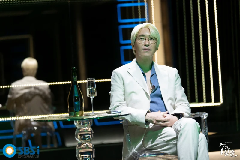 유리로된 테이블에 고급 와인이 있는 고급바에 흰 양복과 금발에 안경을 쓴 드라마 7인의 탈출의 매튜 리 캐릭터