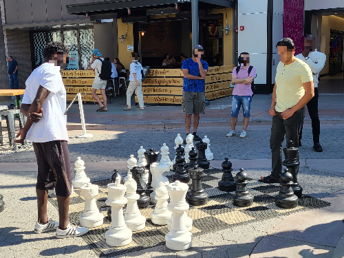 거리에서 큰 말로 체스를 두는 사람들의 모습. 체스의 말은 성인 남자의 종아리 길이보다 좀 더 큰 크기이다. 두 명이 마주 보며 체스를 두고 있고&amp;#44; 구경하는 사람들이 있다.