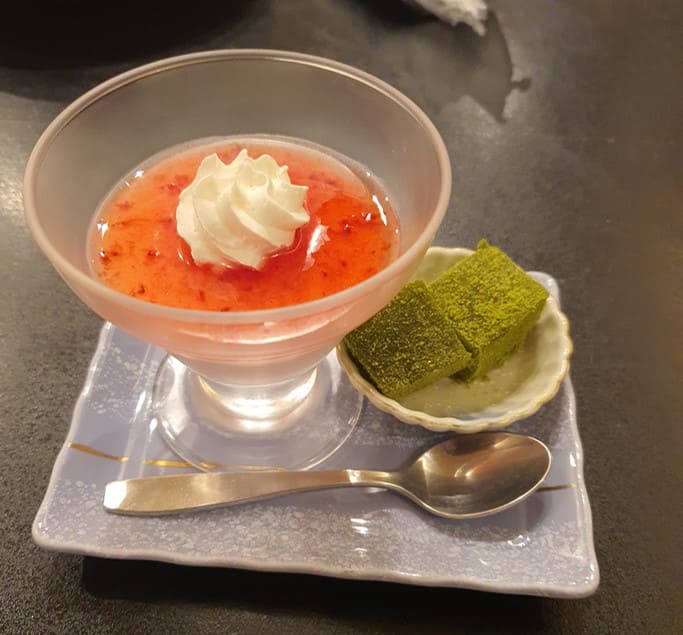 투명한 그릇안에 빨간색 푸딩 그리고 그 위에 크림이 올라가 있고 옆으로 작은 접시에 녹색의 떡이 있다.