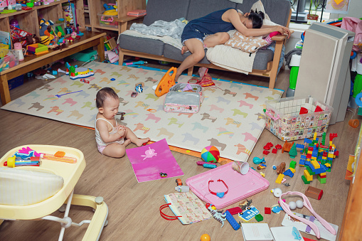 잔뜩 장난감이 어질러진 거실에서 아기가 혼자 놀고있고 엄마는 소파에 엎드려 피곤해하는 사진