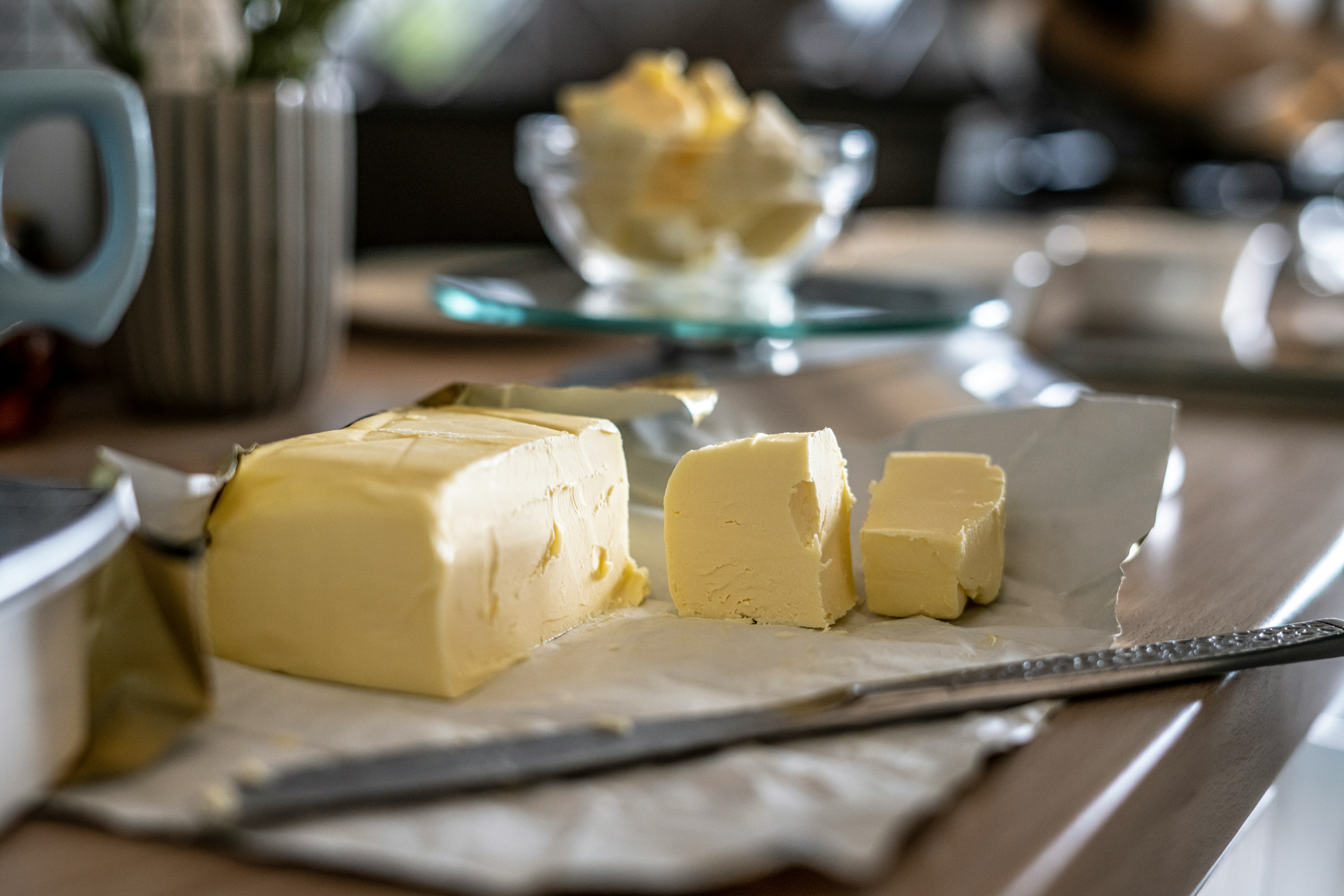 고동색의 네모난 테이블 위에 버터의 껍질을 까서 칼로 3토박 잘라 놓은 것과 뒤로는 투명한 유리 그릇에 버터를 잘게 잘라서 가득 담아 놓은 것을 두고 찍은 사진