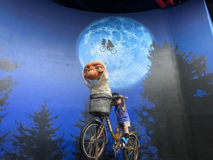E.T.영화에서 주인공과 외계인이 달 앞을 날아가는 배경이 있고 앞에 바구니에 외계인이 앉아 있고 자전거를 타고 있는 작은 아이가 있다.