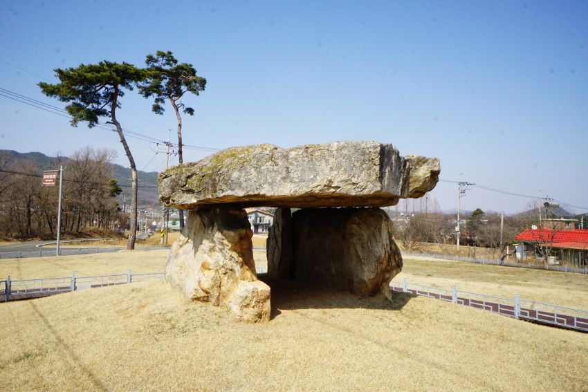 가운데 고인돌 근접시진&#44; 뒤로 키 큰 소나무 둘&#44; 우측에 붉은 지붕의 건물&#44; 푸른 하늘&#44;