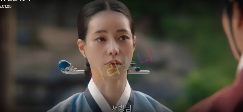 푸른 전통 한복을 입고 머리를 단정히 묶은 태영이 상념에 잠긴 듯한 표정으로 상대를 바라보고 있습니다. 그녀의 뒤로는 흐릿한 돌담과 나무들이 보입니다. 자막은 &amp;quot;서방님.&amp;quot;