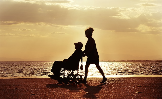 여자가 휠체어 탄 남성 밀고 있는 사진 배경 바닷가