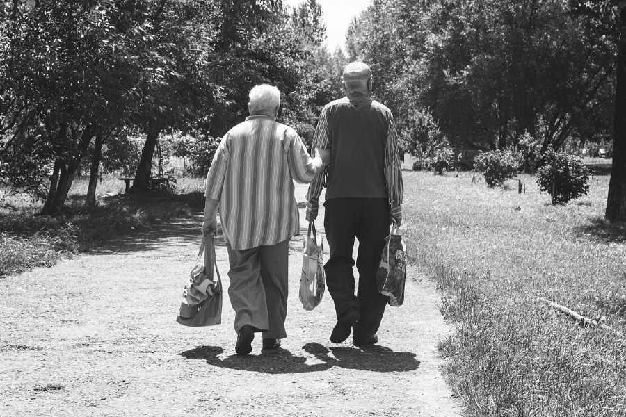 노인 부부가 양쪽 손에 짐을 들고 나란히 한적한 마을 길을 다정하게 걷고 있는 모습을 흑백으로 찍은 사진