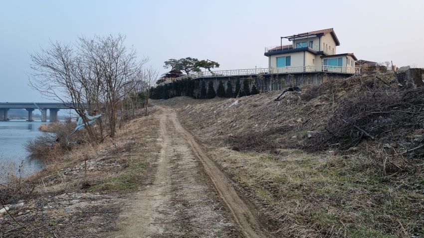 강변에 흙길, 우측에 보이는 2층집, 흰벽, 파란 유리창, 집에 둘러쳐진 큰크리트 담벼락, 소나무 몇그루 왼쪽 멀리 보이는 대교, 흐린 하늘,