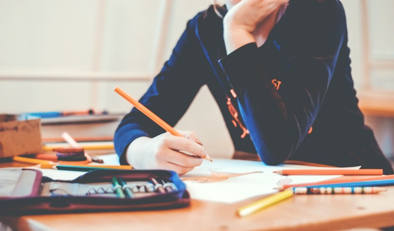 A student sitting at a desk surrounded by colored pencils and writing in a notebook, symbolizing academic preparation for post-graduation career planning.