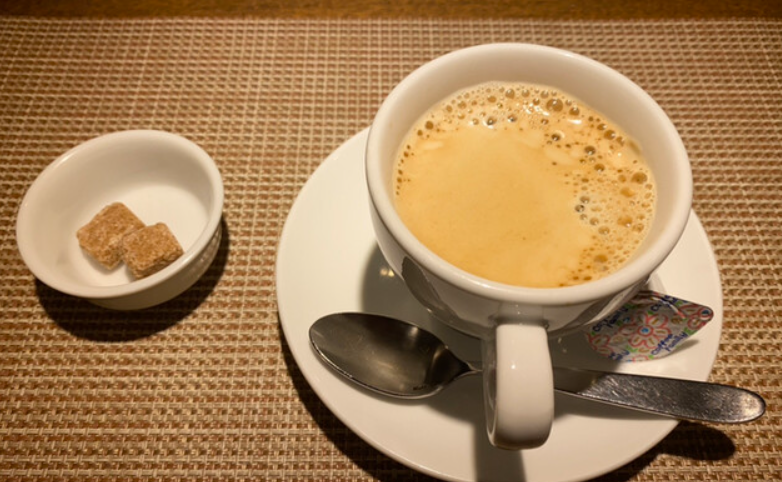 테이블 위에 흰색의 커피잔에 커피가 있고 조그마한 종지에 설탕이 올려져 있다.
