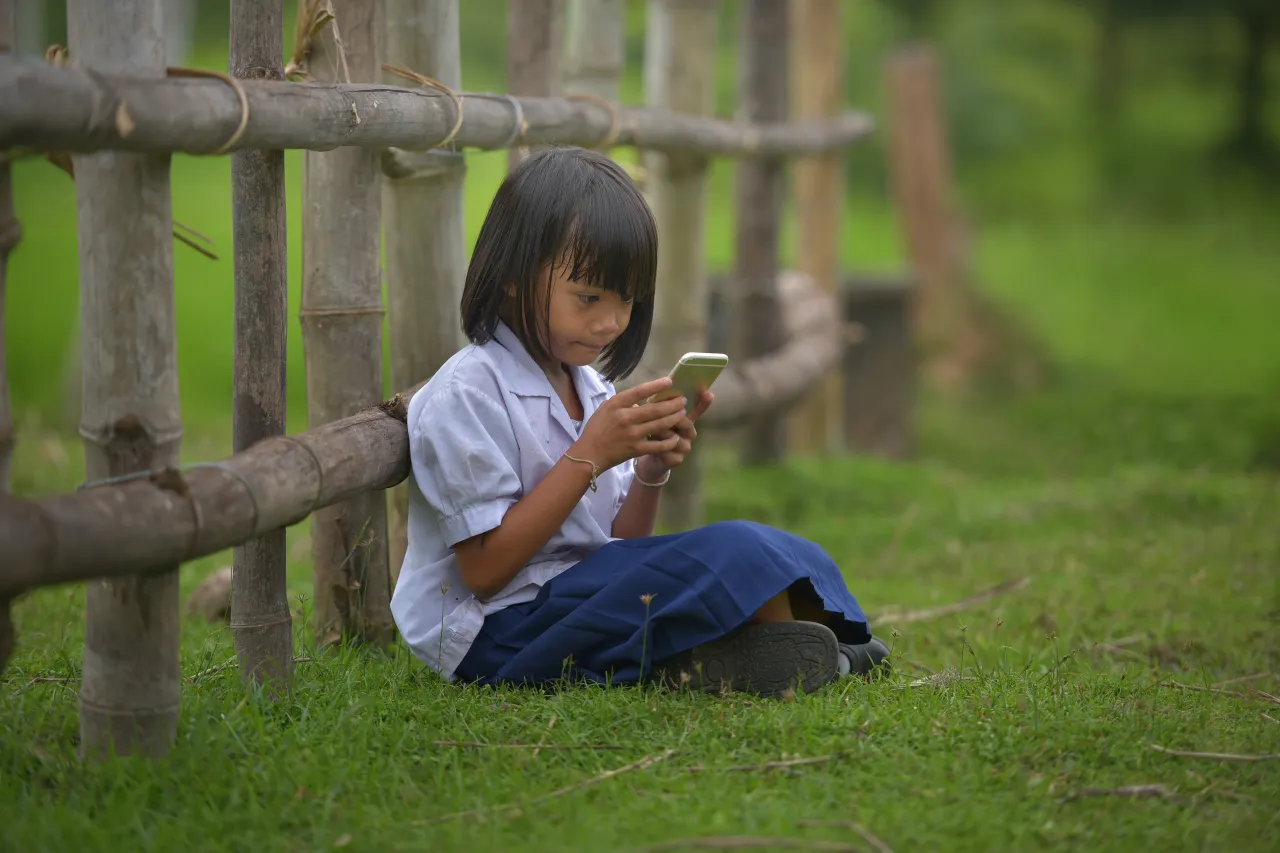 캄보디아 여자 어린이가 울타리에 기대어 앉아 스마트폰을 하고 있는 모습