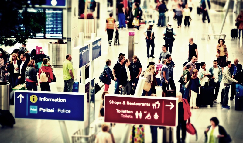 A crowded international airport terminal with travelers and directional signs for information, police, toilets, shops, and restaurants.