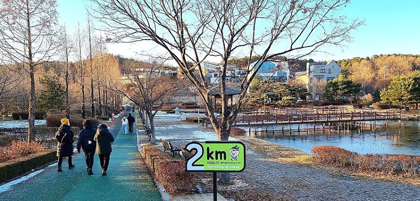 왼쪽 녹색 산책로에 여성 3, 뒷모습, 그 앞에 커플 1쌍, 뒷모습, 왼쪽에 늘어선 메타세콰이어, 가운데 꽂힌 연두색 팻말에 표시돤 2km, 우측에 연못, 연못 위에 탐방로, 푸른하늘,