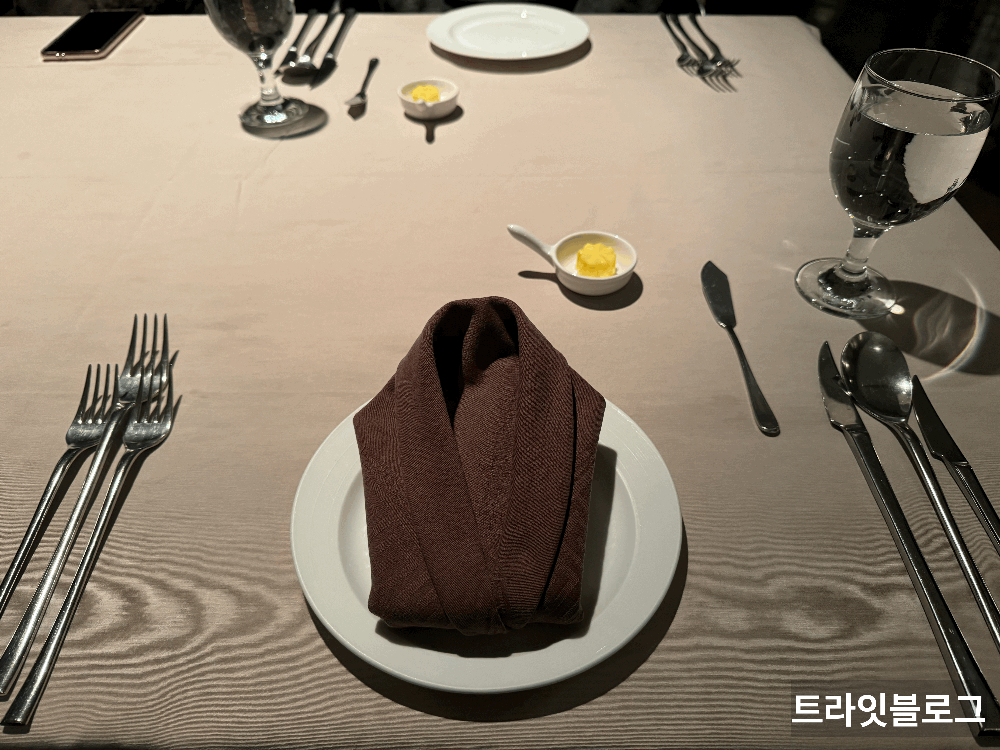 테이블
접시 위에 냅킨이 접혀져 있고
접시 왼쪽엔 포크 세개
접시 오른쪽엔 나이프 2개&#44; 숟가락 1개가 있다.