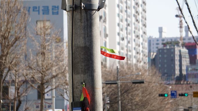 가로등에 매어져 바람에 나부끼는 삘긴 리본&#44; 뒤로는 아파트 건물&#44; 신호등&#44; 직진 파란불&#44;