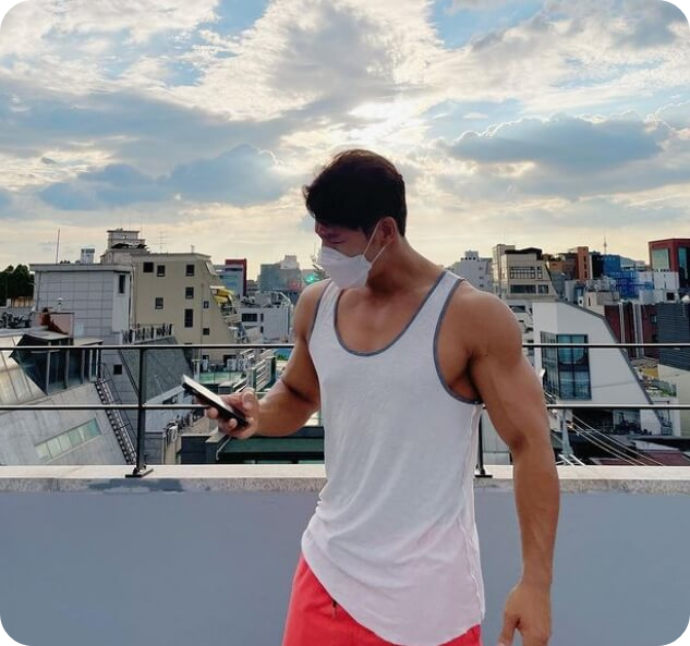 옥상 위에서 구름 낀 하늘과 도시를 배경으로 찍은 김종국 사진. 흰색 메리야스를 입고 팔 근육을 드러내고 있으며&#44; 휴대폰을 보고 있다.