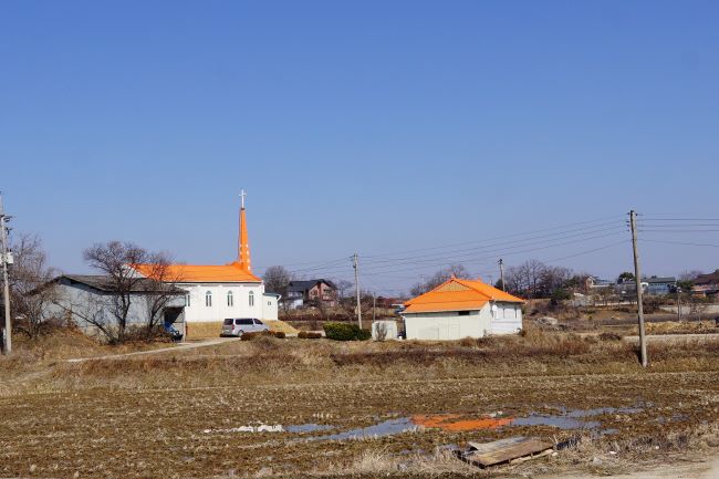 푸른 하늘&#44; 마을 풍경&#44; 오렌지 지붕의 교회 건물&#44; 앞에는 물에 젖은 밭&#44; 우측에 전봇대 1&#44;