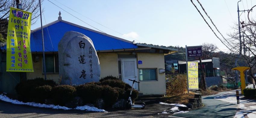 백련암 표지석, 둥근 바위에 한자 글씨, 왼쪽에 노란 세로 현수막에 백련암 100기도 안내문, 뒤에 단층 건물, 파란 지붕, 푸른 하늘, 역광,