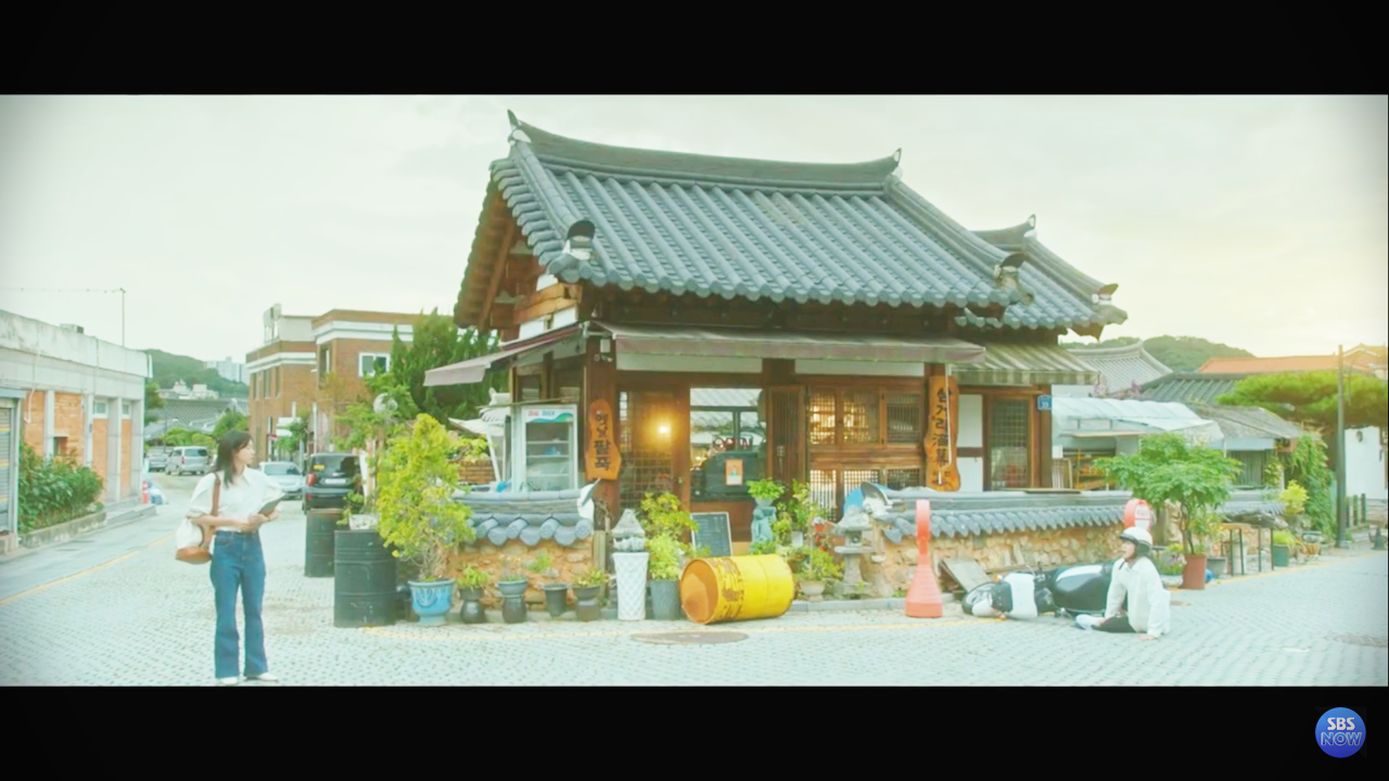 김다미가 왼쪽 도로에 서 있고, 최우식은 오른쪽에서 작은 오토바이를 타다가 자빠져있는 장면이 코믹하게 그려진 사진입니다