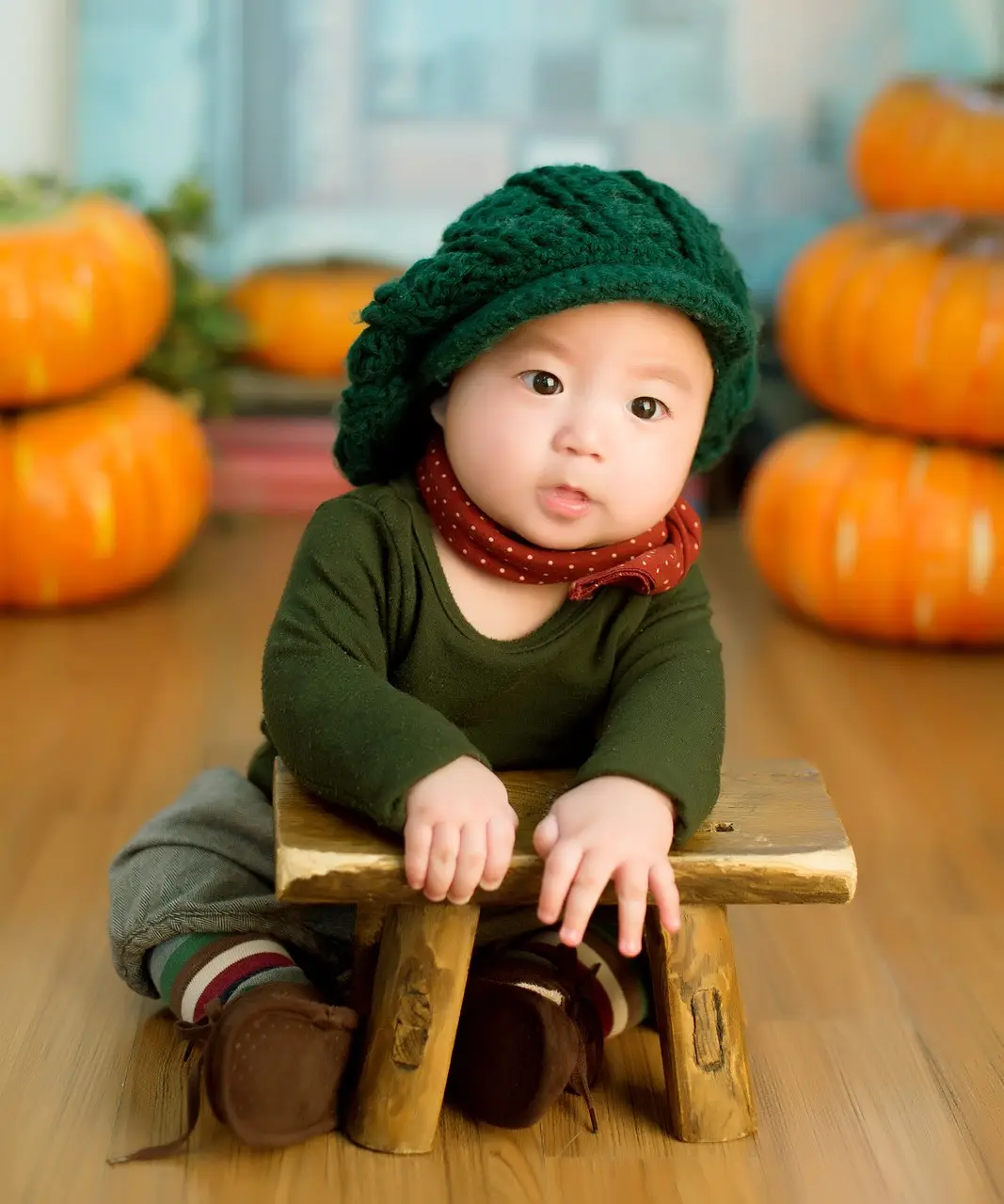 부모급여- 실내 나무바닥위 좌측 우측 호박이 2개씩 쌓인 배경으로 초록색 옷과 모자를 쓰고 앉아있는 아기