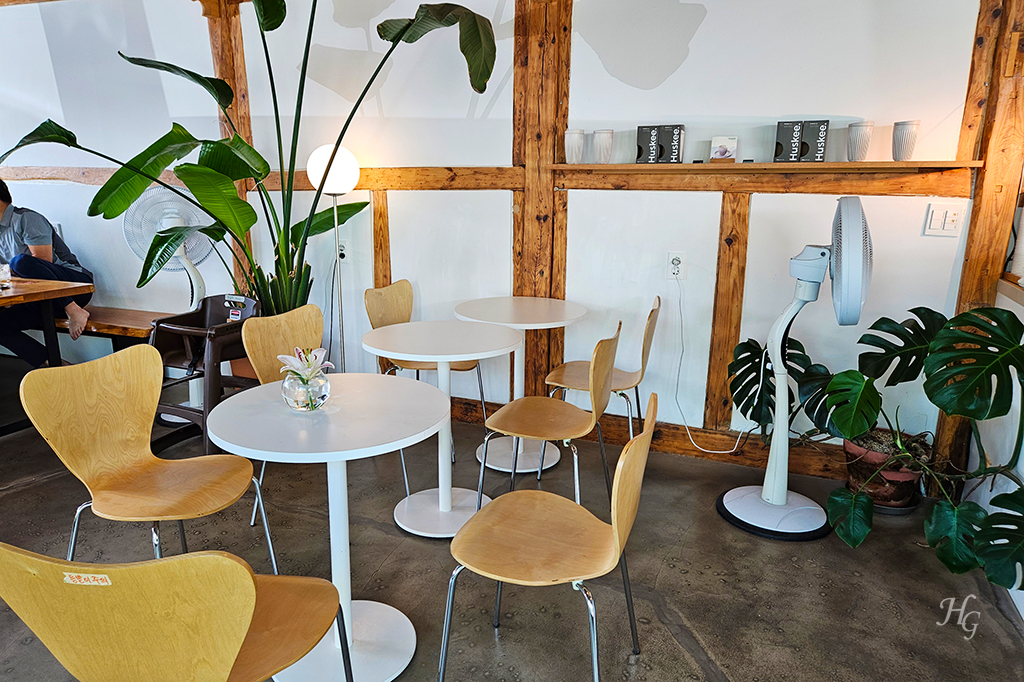 목조 기둥과 식물&#44; 하얀 벽&#44; 하얀 테이블과 나무색 의자 등이 잘 어울리는 진저보이해미 카페 내부