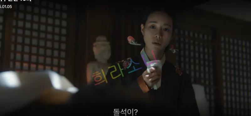 꽃무늬 한복을 입고 머리를 단정히 올린 태영이 하얀 천으로 코를 막으며 깜짝 놀란 표정을 짓고 있습니다. 그녀의 배경에는 불상이 놓여 있습니다. 자막은 &amp;quot;돌석이?&amp;quot;