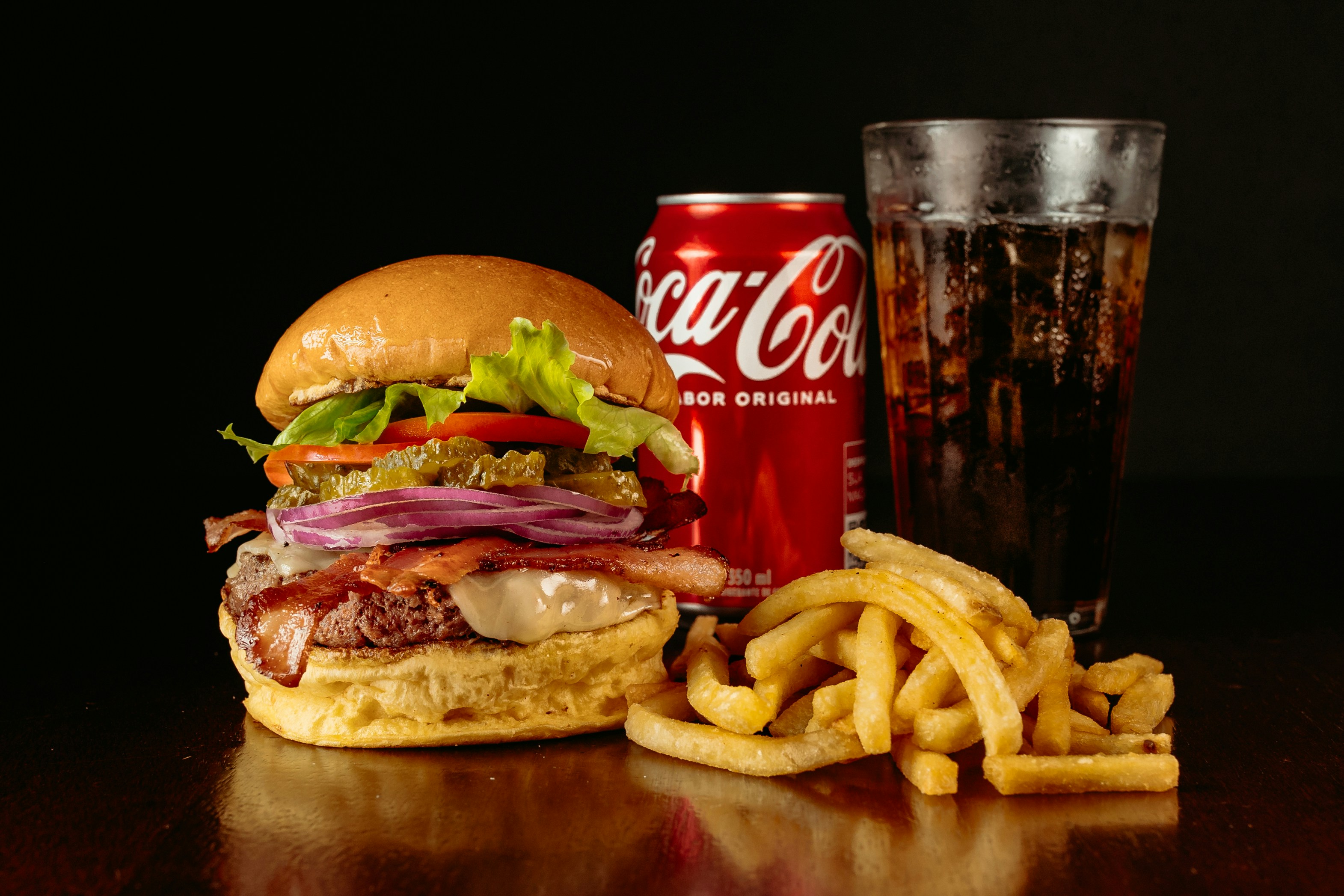 검은 바닥 위에 햄버거와 콜라 캔&#44; 콜라를 가득 따라 놓은 유리컵&#44; 그리고 그 앞에 감자튀김을 놓고 찍은 사진