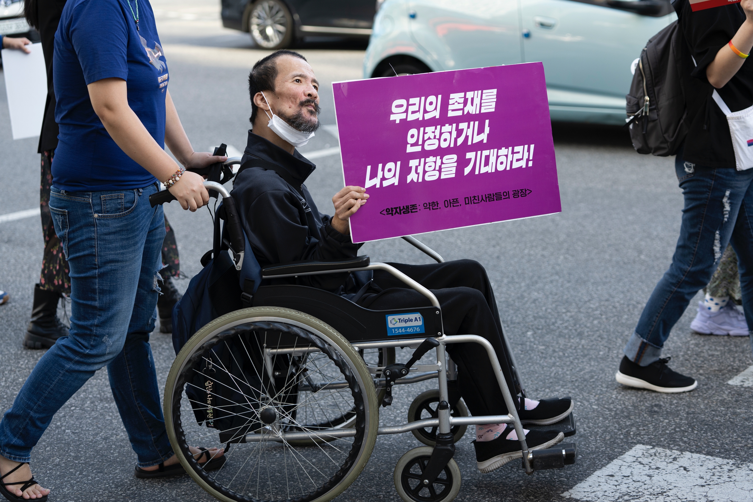 휠체어를 탄 시민이 &#39;우리의 존재를 인정하거나 나의 저항을 기대하라!&#39;라는 피켓을 들고 행진하고 있다.